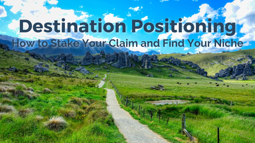 a landscape in New Zealand with bright green grass in the foreground, split by a walking path. In the background there are mountains and a vibrant blue sky with clouds. The title of the blog appears on top of the image: Destination Positioning, how to stake your claim and find your niche